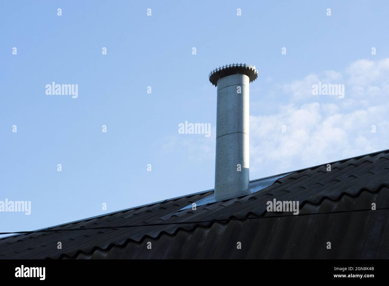 Galvanized iron furnace tube on old dark tile roof on a light haze clouds on blue sky Stock Photo