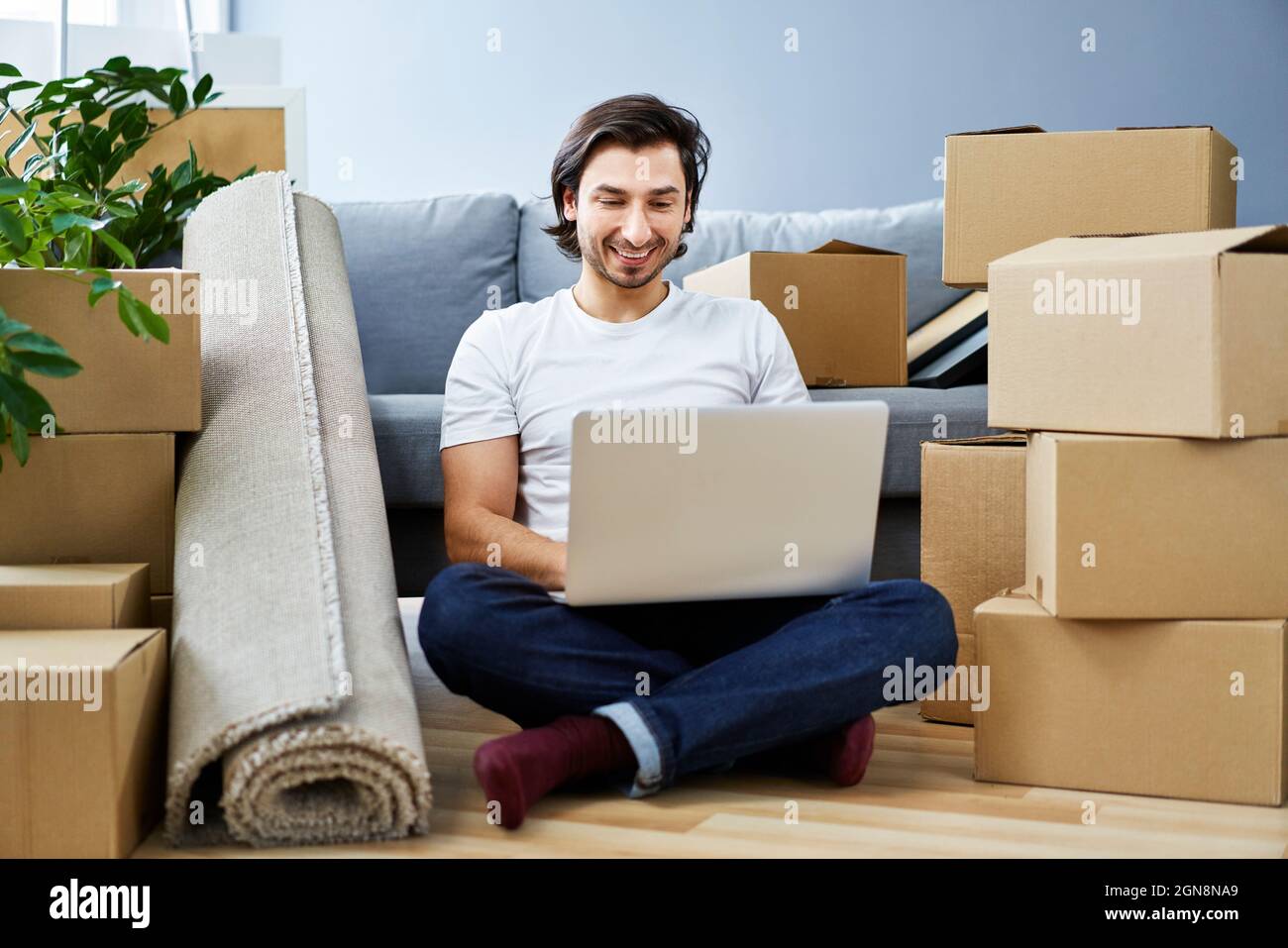 Smiling man using laptop while sitting amidst cardboard boxes at home Stock Photo