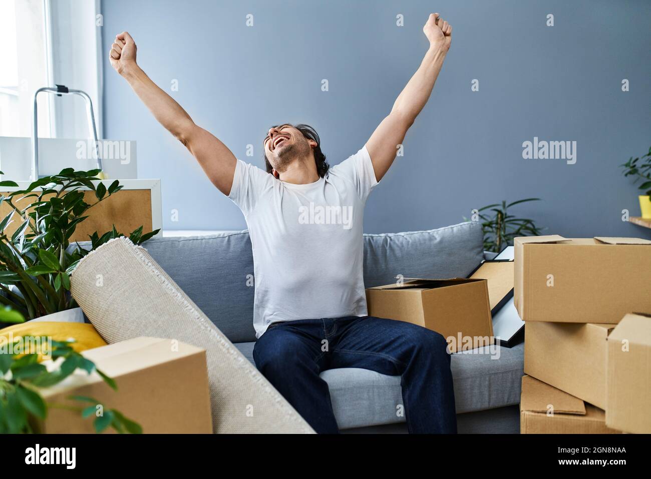 Happy man with arms raised sitting on sofa amidst cardboard boxes at ...