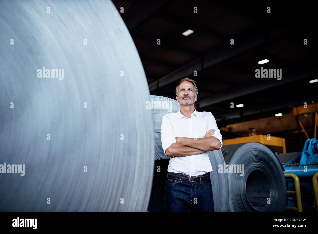 Thoughtful businessman with arms crossed standing by steel sheet roll Stock Photo