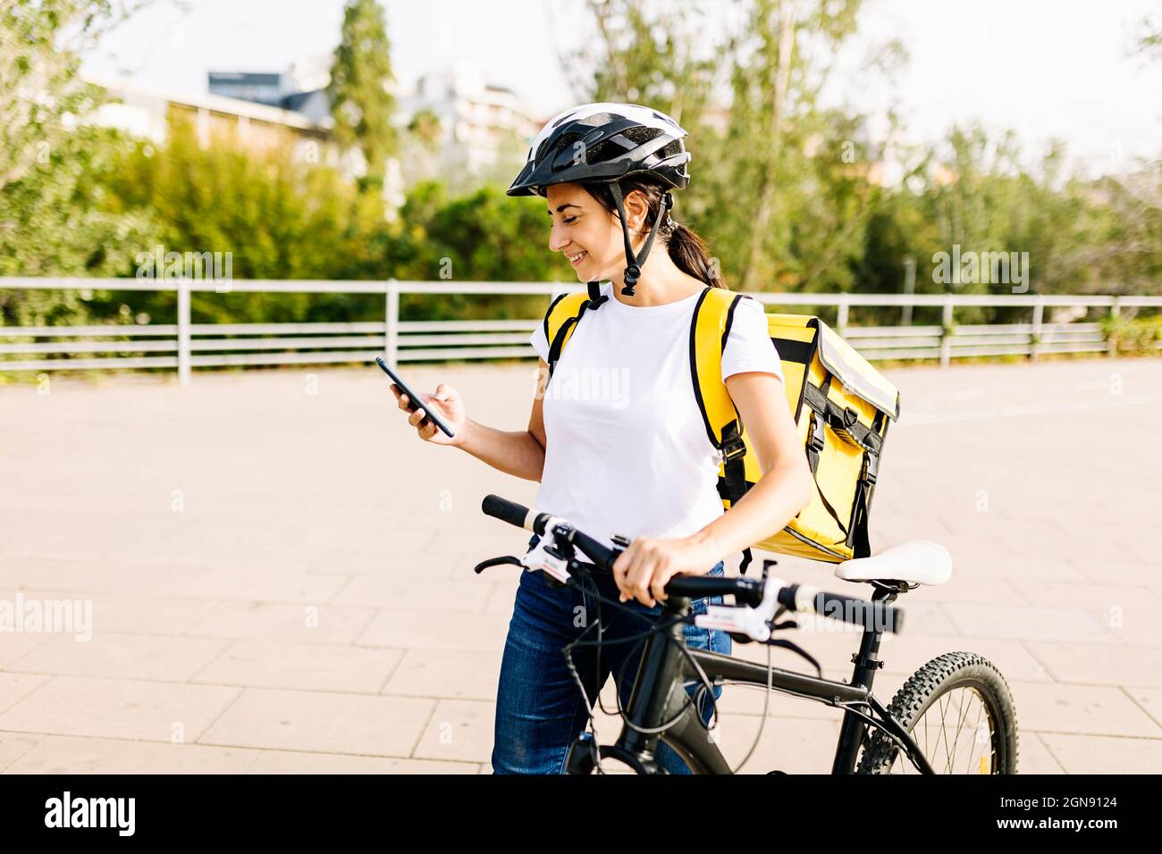 Smiling delivery woman using mobile phone while wheeling bicycle Stock Photo