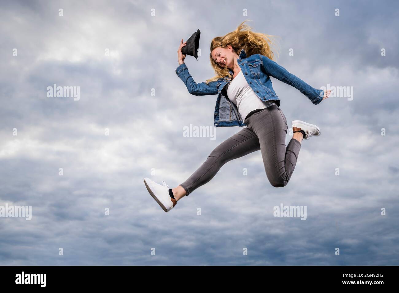 Young woman jumping against cloudy sky with hat in hand Stock Photo