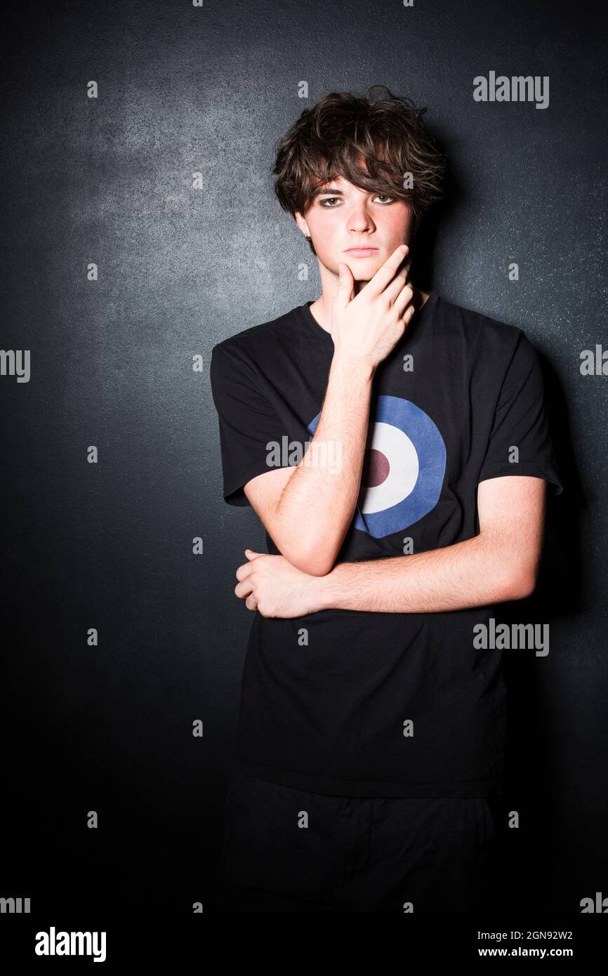 Male teenager standing with hand on chin against black color background Stock Photo