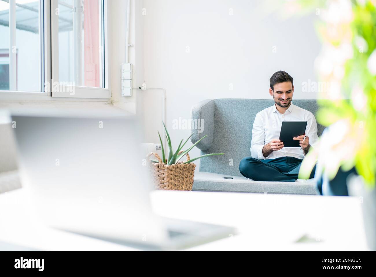 Smiling young businessman using digital tablet while sitting on sofa in office Stock Photo