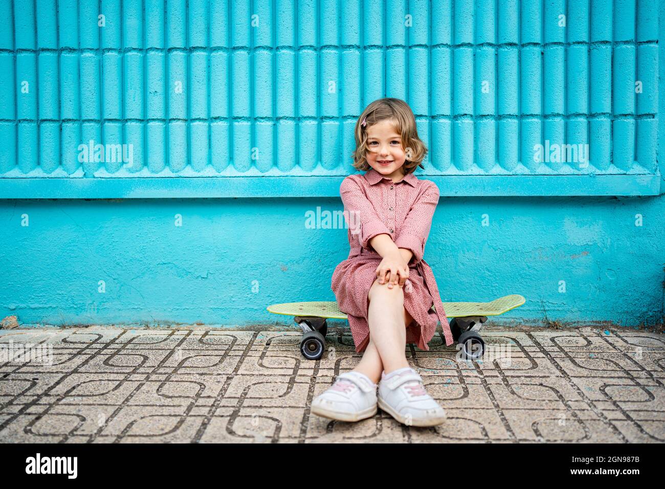 Smiling girl sitting with legs crossed at knee on skateboard in front of blue wall Stock Photo