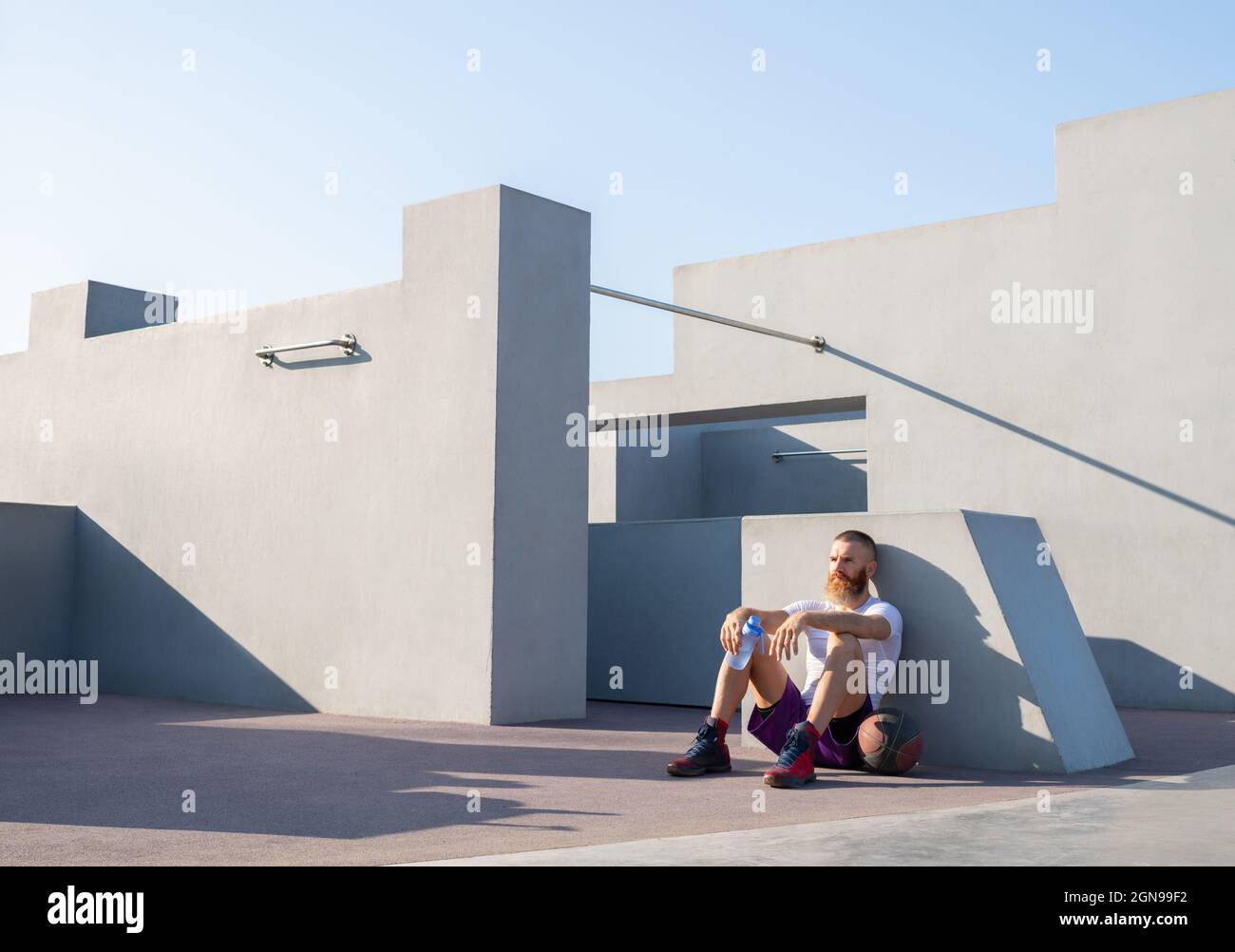 Tired adult bearded basketball player sitting near concrete wall and relaxing with bottle of water Stock Photo