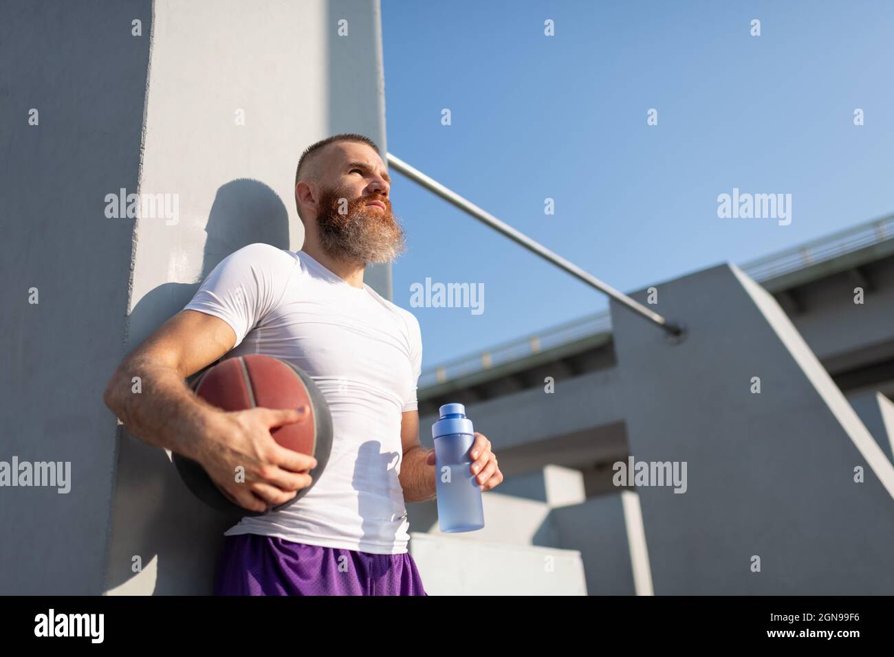 Determined bearded athlete with basketball ball and bottle of water looking away while taking break Stock Photo