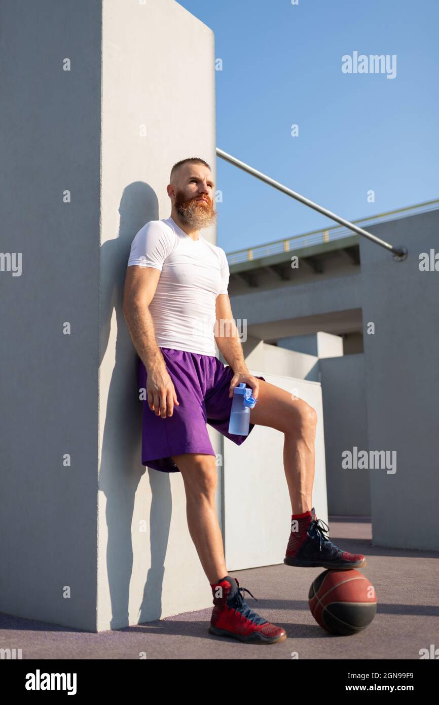 Brutal sportsman in activewear stepping on basketball ball while resting near concrete wall Stock Photo