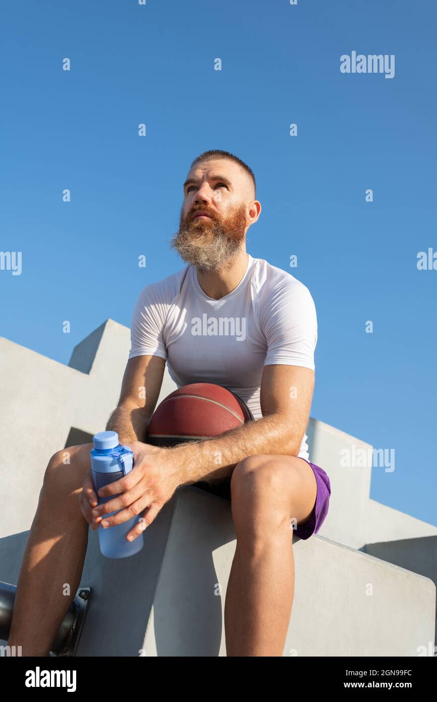 Serious adult sportsman with ball and bottle looking away while sitting on concrete steps Stock Photo