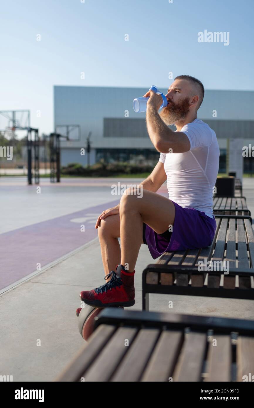 Bearded athlete with ball sitting on bench and sipping water on streetball court Stock Photo