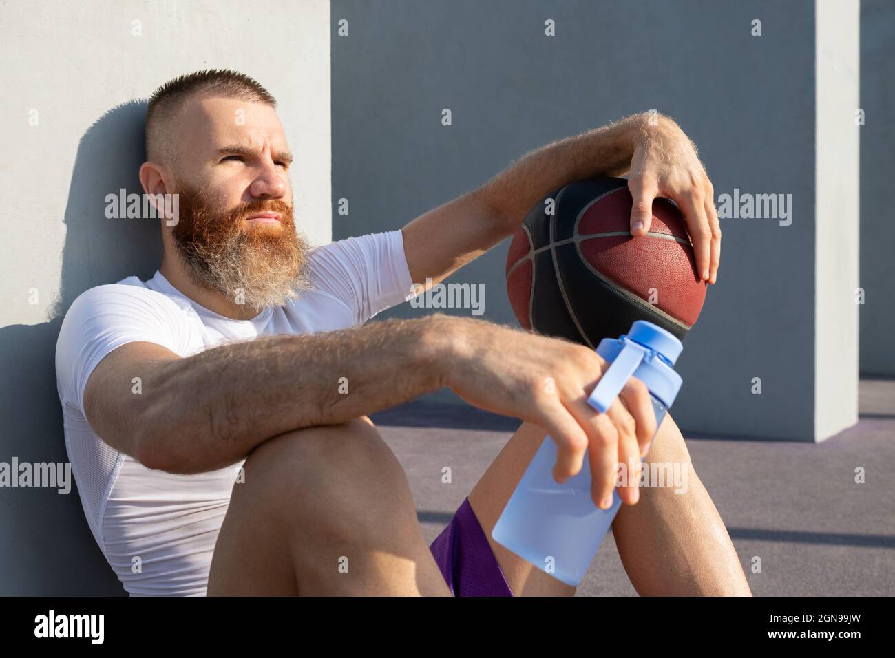 Sportsman with basketball ball and bottle of water looking away thoughtfully while relaxing after workout Stock Photo