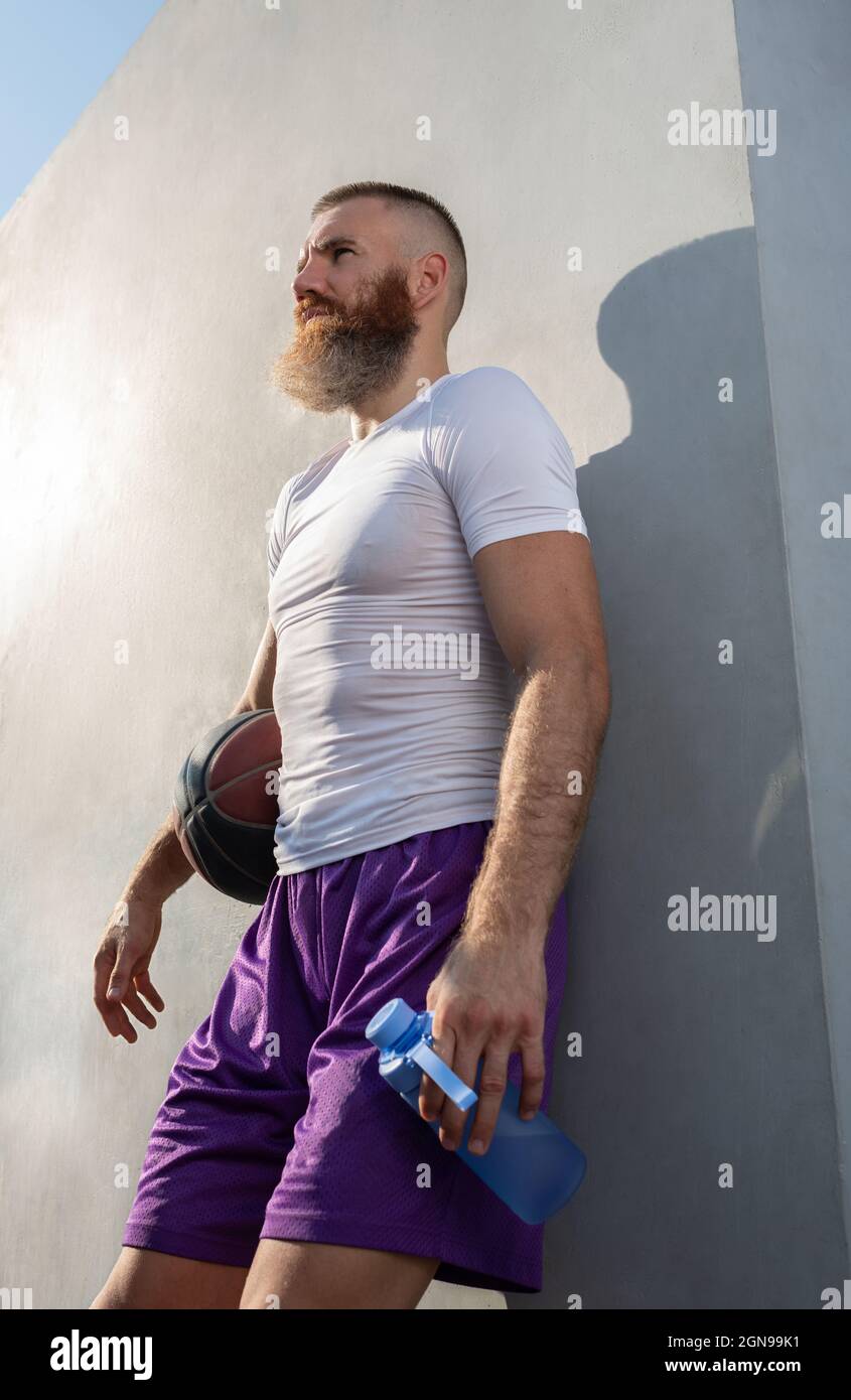 Fit bearded athletic adult sportsman leaning against gray wall while resting after basketball training Stock Photo