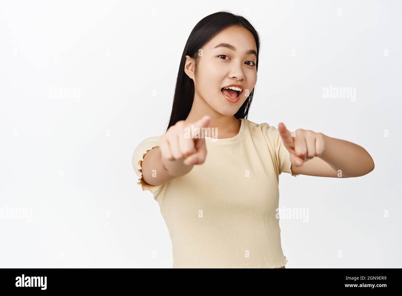 Close up portrait of brunette smiling girl, pointing fingers at camera, choosing you, inviting to event, standing in t-shirt over white background Stock Photo
