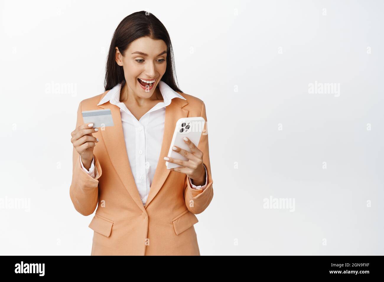 Saleswoman looks excited at smartphone screen app while placing order online store, holding credit card and phone, standing over white background Stock Photo