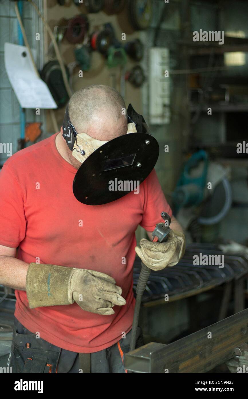 A masked welder works in a workshop. The man works with metal. The guy works in the workshop. Welding of steel seam. Stock Photo
