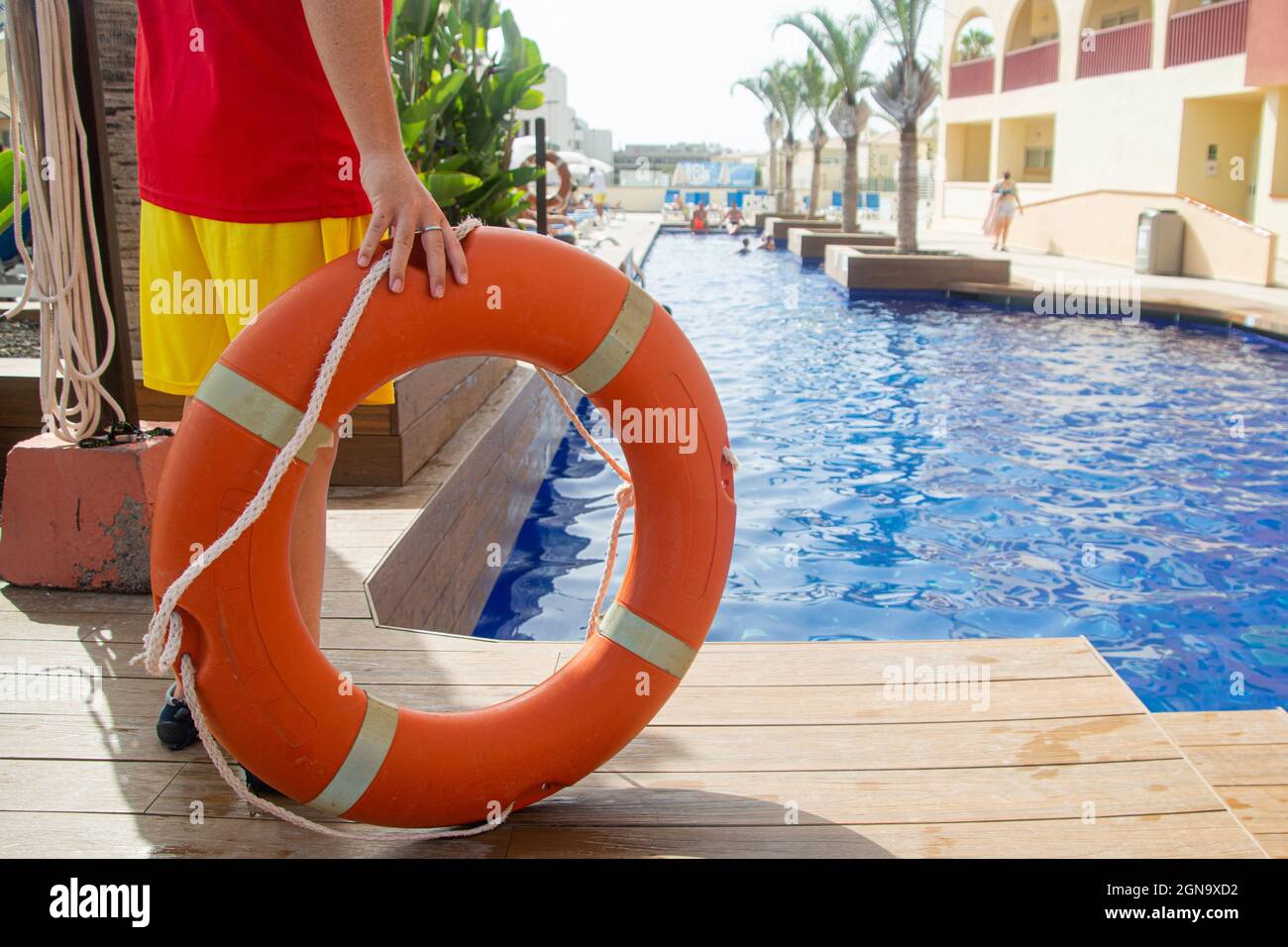 Close up of a lifebuoy ring with rope held up by a lifeguard with a swimming pool on the background . Lifeguard during work concept Stock Photo