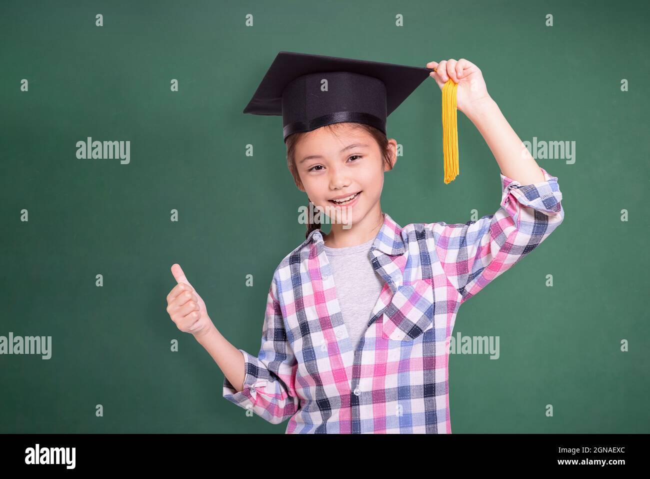 Happy student girl wearing graduation cap and showing thumb up.Isolated on green chalkboard background. Stock Photo