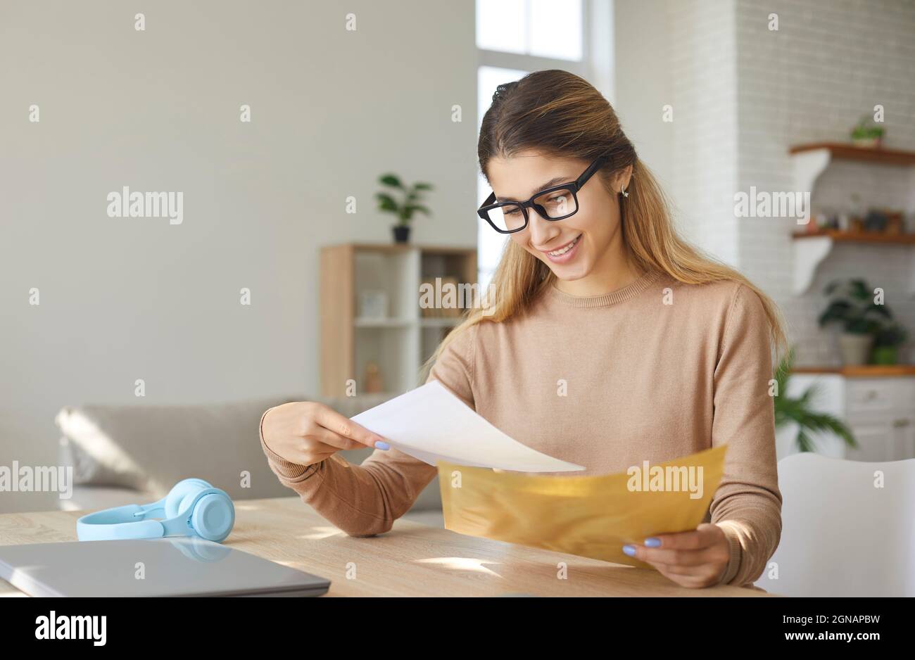 Smiling young woman who is excited by good news reads letter unfolded from paper envelope. Stock Photo