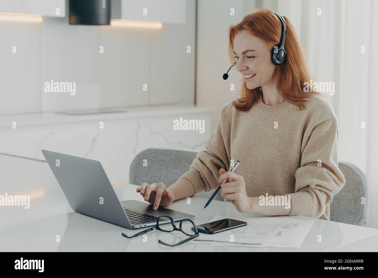 Happy redhead woman watches online webinar uses headset focused at laptop screen Stock Photo