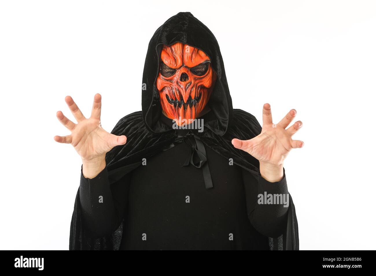 Man dressed in pumpkin latex mask and hooded velvet cape, scares with his hands, on white background. Carnival, Halloween and days of the dead concept Stock Photo