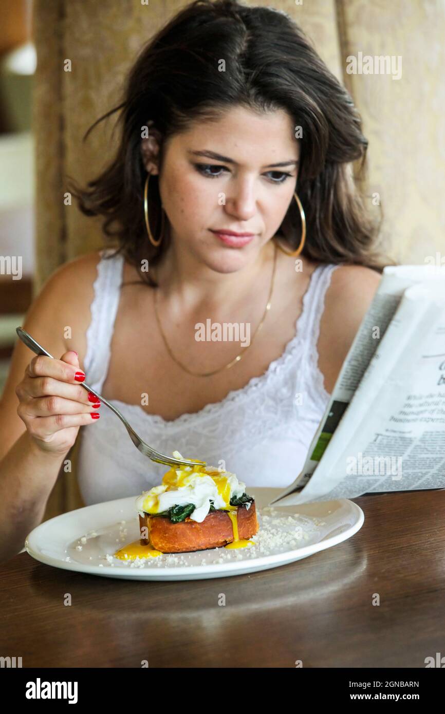 Young woman eats Eggs Benedict while reading the morning newspaper Stock Photo