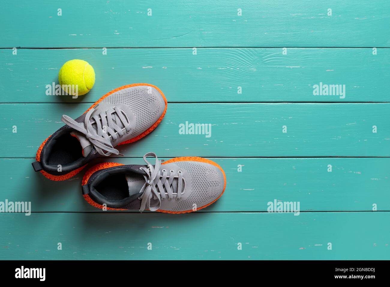 Laced up gray textile sneakers and tennis ball over turquoise wood background. Pair of new mesh fabric trainers with orange sole. Stylish sport shoes. Stock Photo