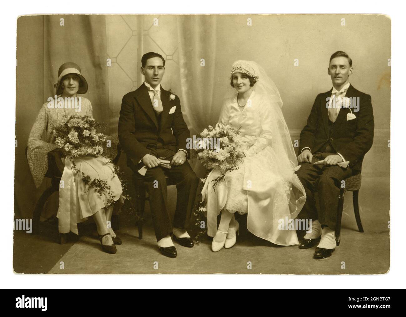 Stunning original classic 1920's, flapper era, wedding group portrait photo, beautiful bride wearing a long veil, stunning headpiece. sitting next to the groom and best man wearing matching suits with winged shirt collars, spats. The bridesmaid or maid of honour is wearing a fashionable cloche hat, circa 1924, U.K. Stock Photo