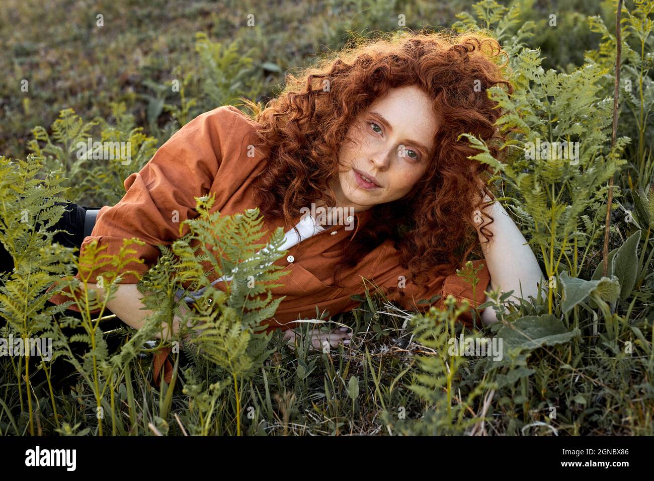redhead woman lying on grass surrounded by fantastic fairy plants, curly lady with natural red hair posing at camera, wearing orange casual shirt, in Stock Photo