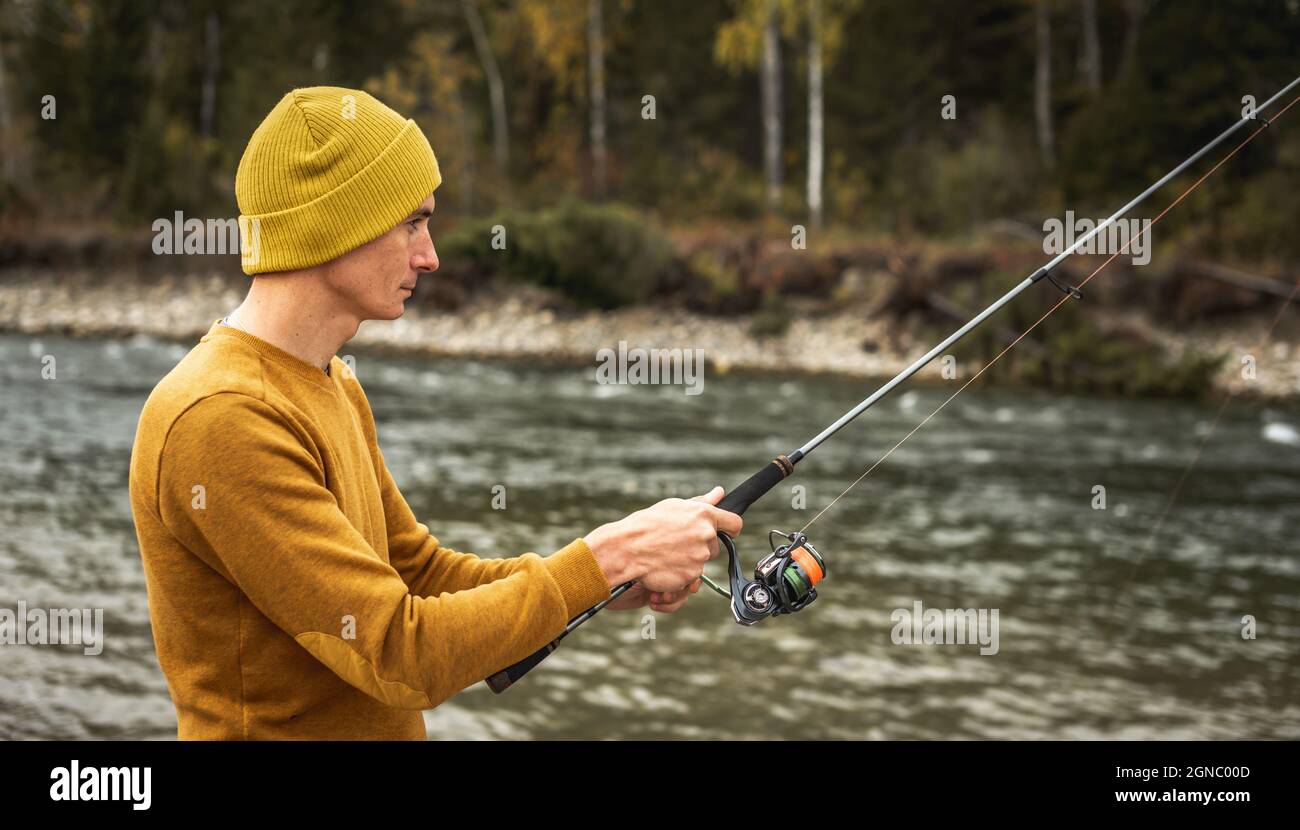 Young man fisherman in a yellow sweater and hat is fishing on the river  bank in the autumn forest with a spinning rod in his hands. Concept of  relaxin Stock Photo 