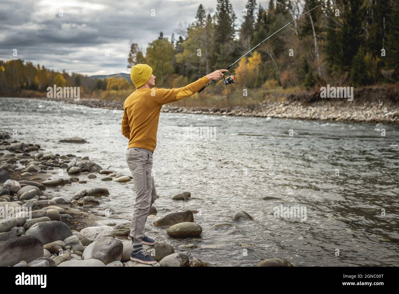 Young man fisherman in a yellow sweater and hat is fishing on the stony  river bank