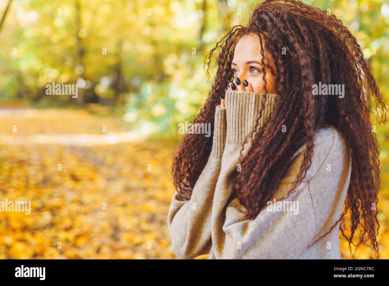 Beautiful afro-haired woman wearing warm sweater walk in autumn park at sunny warm day and feel cold. Portrait of woman outdoor Stock Photo
