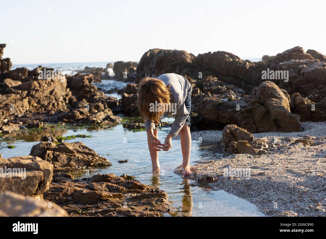 Young boy exploring a rock pool among the jagged rocks of the Atlantic Ocean coastline at sunset, De Kelders, Western Cape, South Africa. Stock Photo
