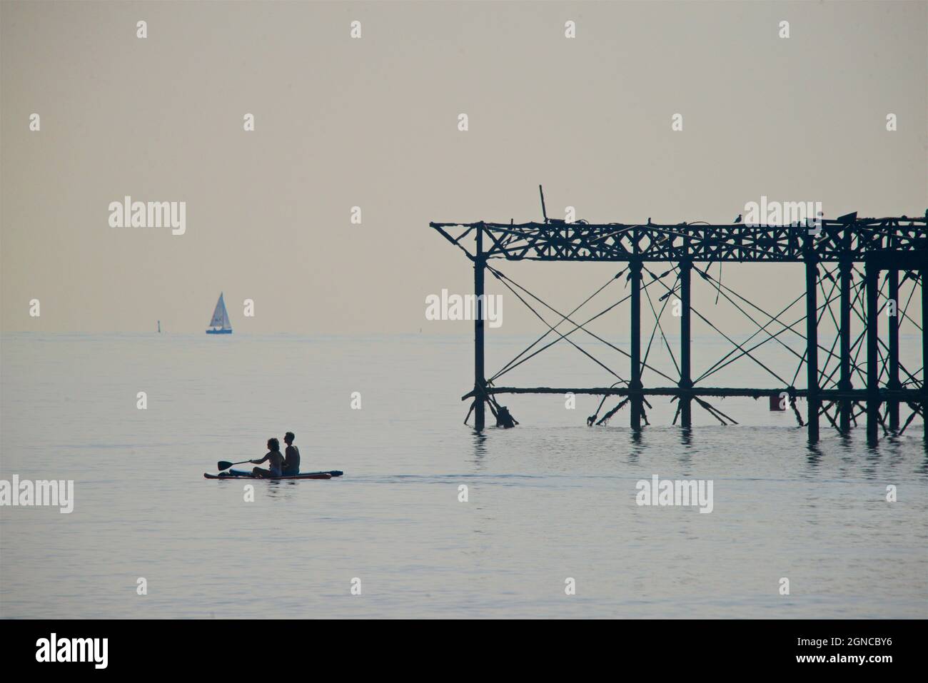 The dilapidated West Pier, Brighton. East Sussex, England. Looking east from Hove.  East Sussex, England. Two paddleboarder ssilhouetted against the skeletal rusting structure of the old pier. Stock Photo