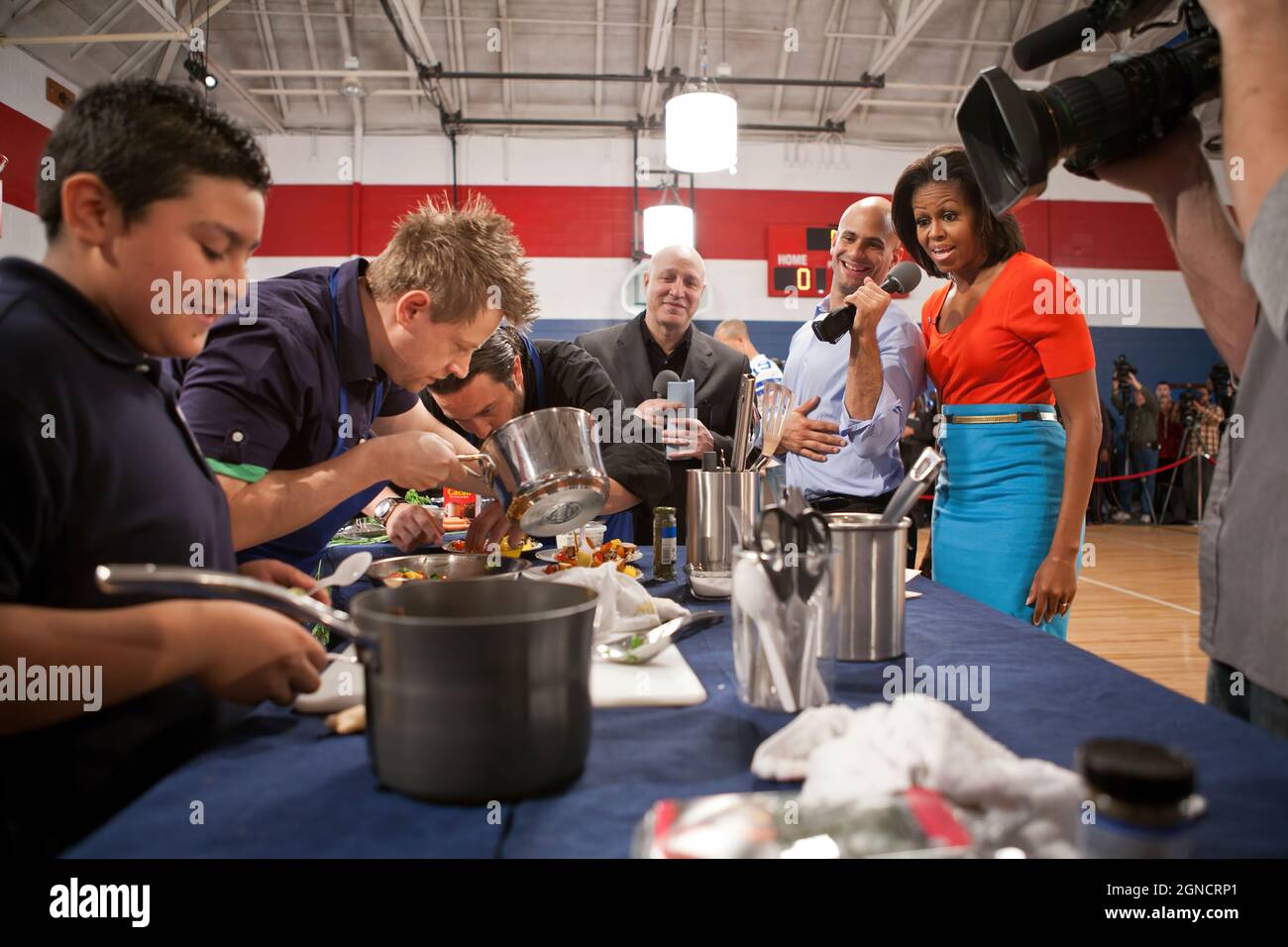First Lady Michelle Obama, Sam Kass, Senior Policy Advisor for Healthy Food Initiatives, and Top Chef head judge Tom Colicchio, center, watch as chefs from past seasons of “Top Chef' and students take part in a cooking competition at the Kleberg Rylie Recreation Center in Dallas, Texas, Feb. 10, 2012. The event, which helped celebrate the second anniversary of the 'Let's Move!' initiative, highlighted the work being done in schools across America to provide healthier food to students. (Official White House Photo by Sonya N. Hebert) This official White House photograph is being made available o Stock Photo