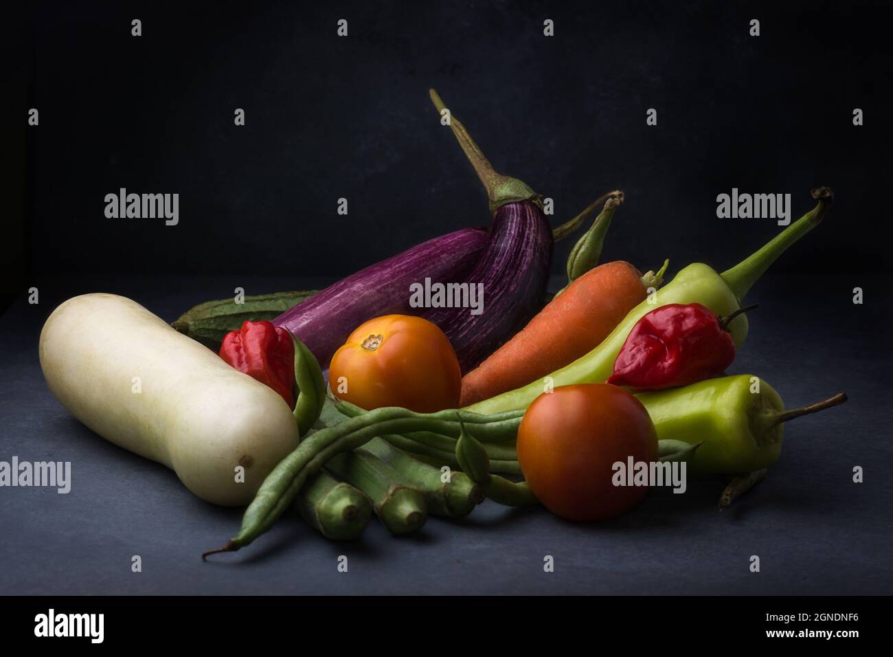 various organic vegetables on a dark textured surface, side view of eggplants, carrot, tomatoes, okra, cucumber and green chilies Stock Photo