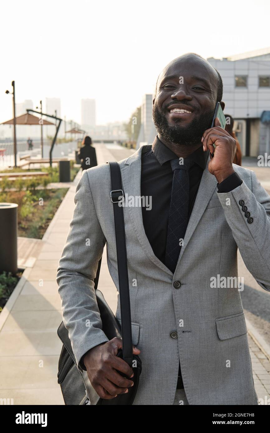 Vertical medium portrait of joyful handsome African American man walking along street after finishing work in office having phone call Stock Photo