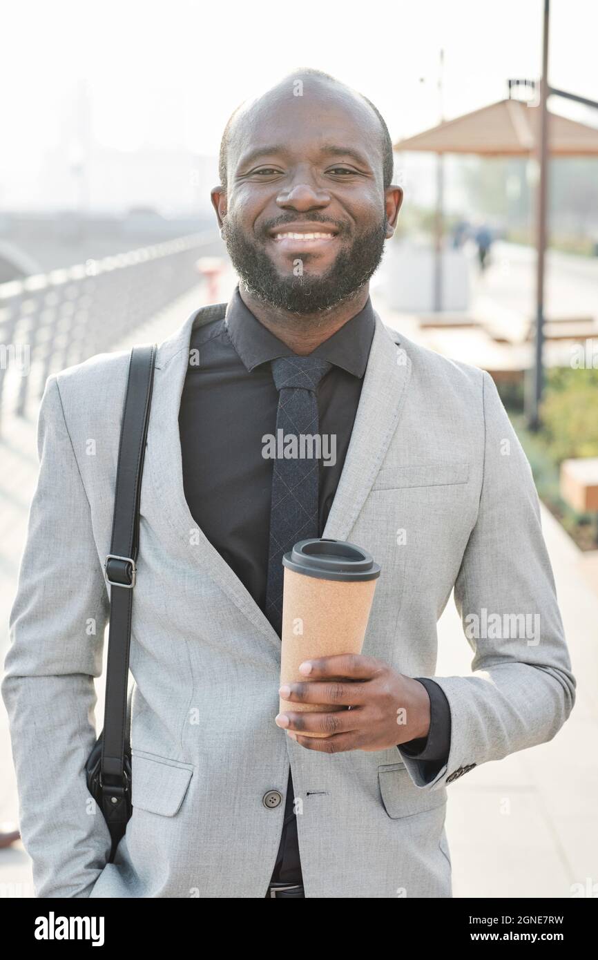 Vertical medium portrait of joyful handsome African American man holding coffee cup spending time outdoors after finishing work in office Stock Photo