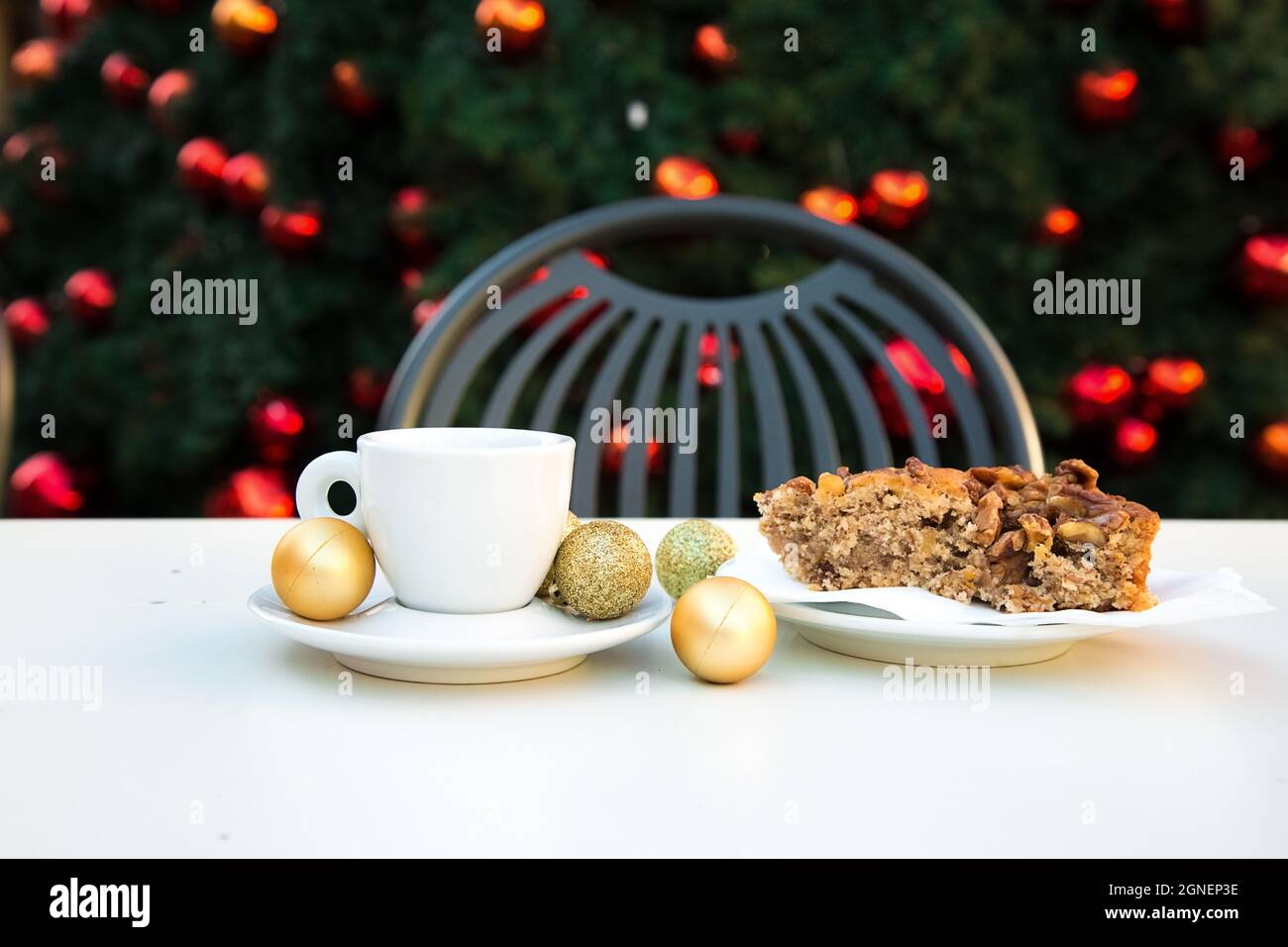 Coffee pair close with golden Christmas balls and a piece of caramel cake with nuts. White table, grey chair and new year tree with red toys in Stock Photo