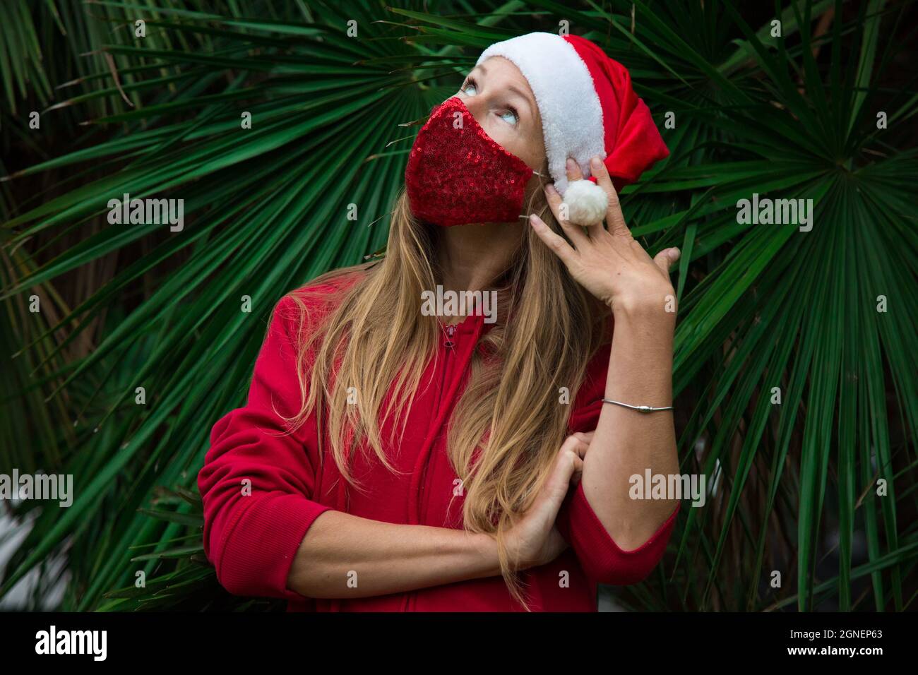 Caucasian blond girl in red hat, coat and red protective face mask with palm leafs behind. Thinking of Christmas 2020. Celebrating pandemic new 2021 Stock Photo