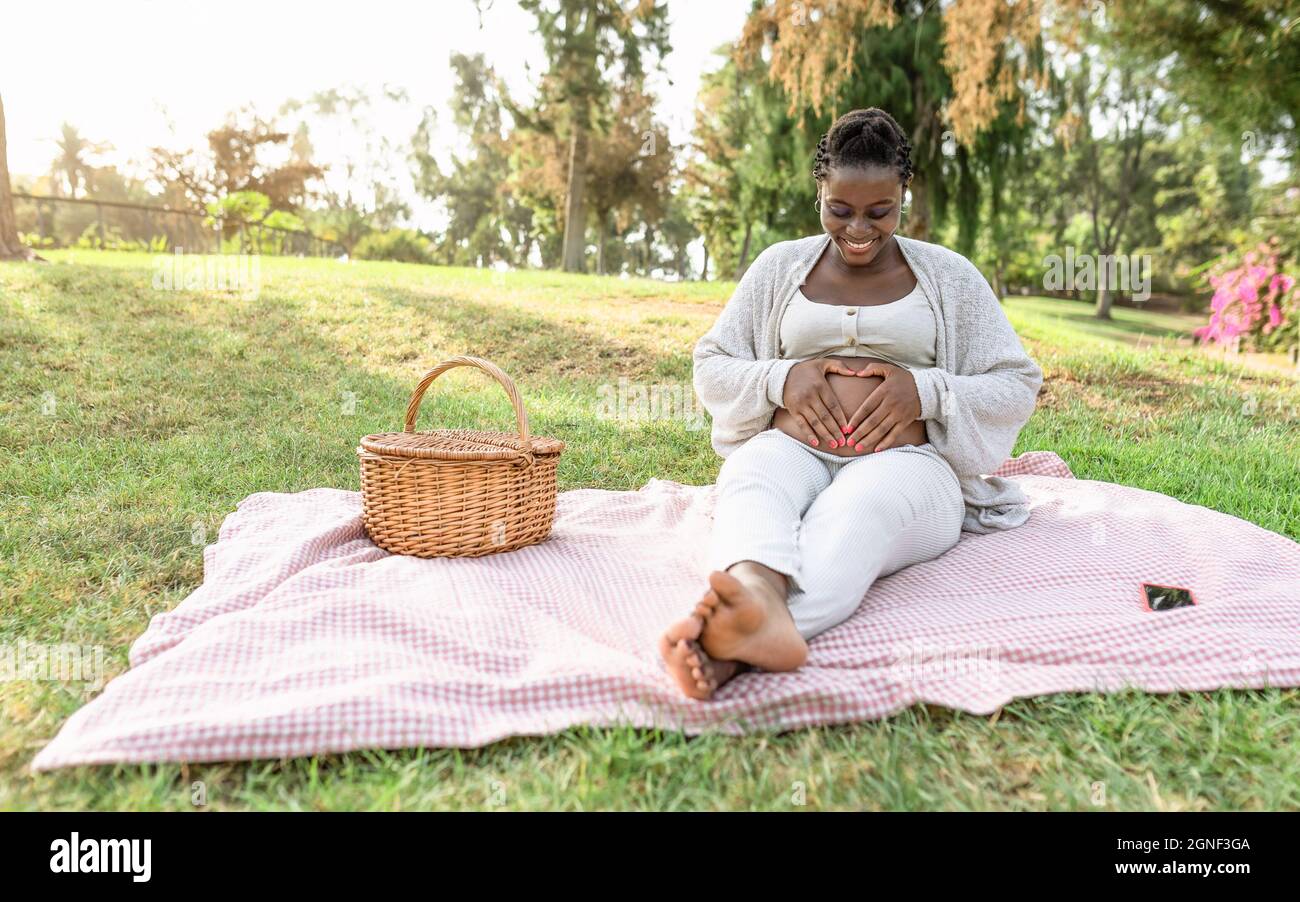 Happy Afro woman touching her pregnant belly doing heart shape with hands in public park - Maternity lifestyle concept Stock Photo