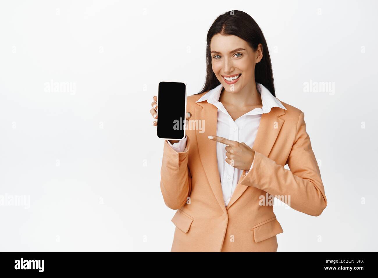Smiling saleswoman pointing finger at mobile phone, recommending online store, showing app interface, standing over white background Stock Photo