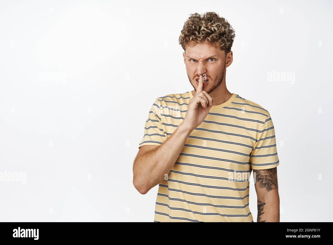 Blond guy grimacing angry and shushing, making hush gesture, telling to be quiet, shut up, dont speak sign, standing over white background Stock Photo