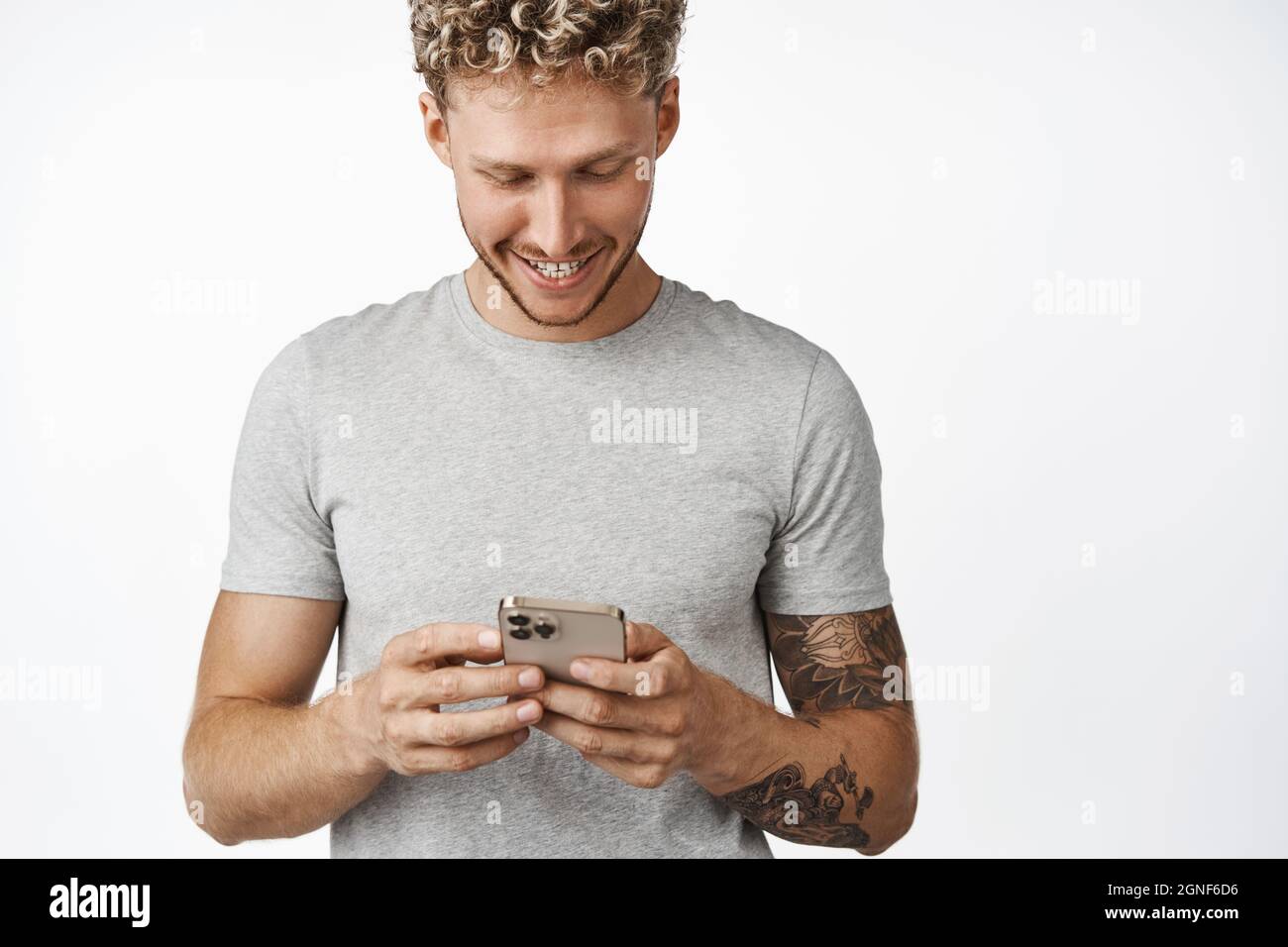 Portrait of handsome blond caucasian man using mobile phone and smiling, looking at his smartphone screen, chatting on cellphone, standing in gray t Stock Photo
