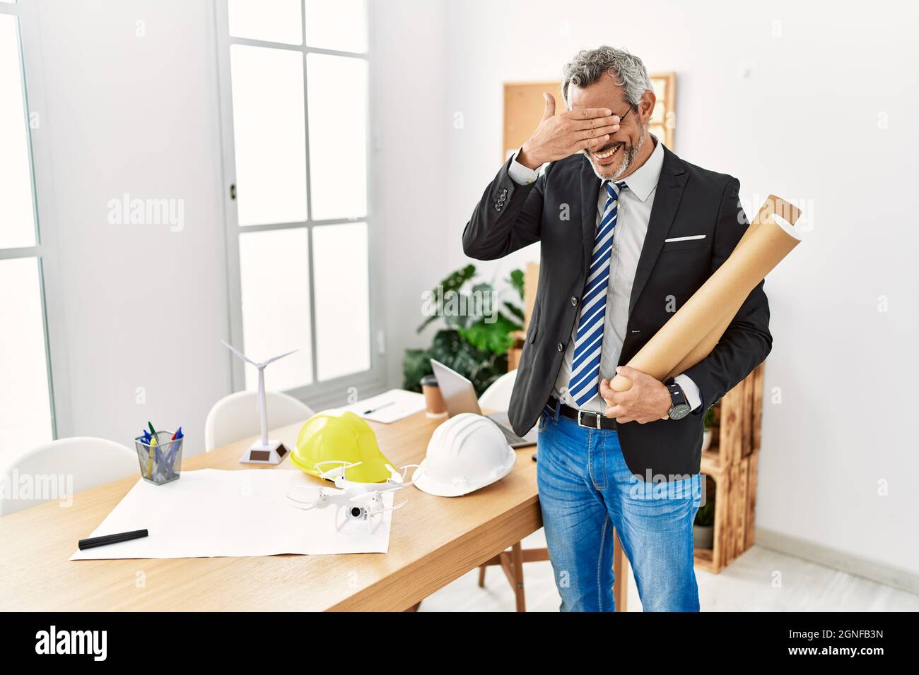 Middle age hispanic business man holding paper blueprints smiling and laughing with hand on face covering eyes for surprise. blind concept. Stock Photo