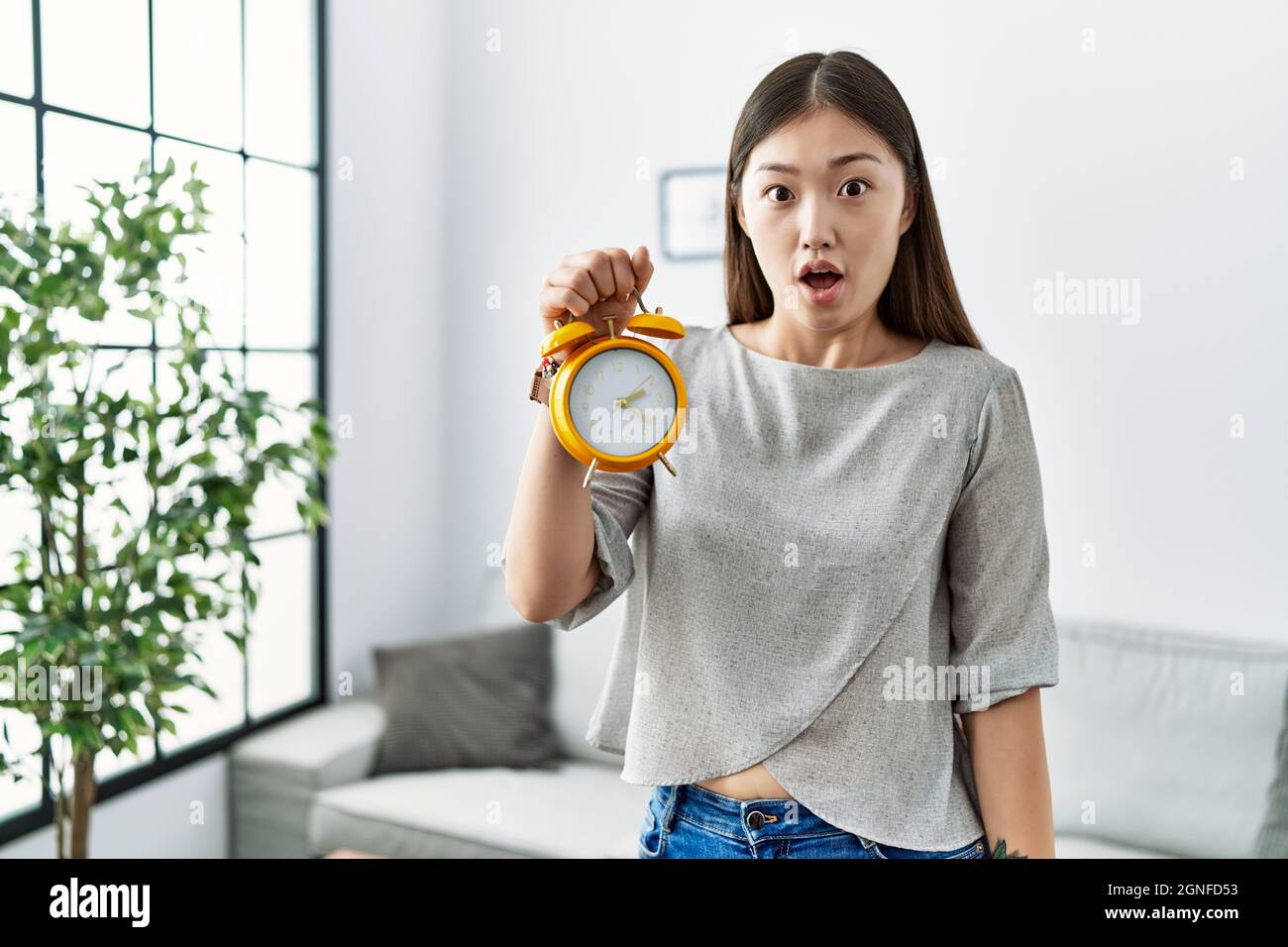 Young asian woman holding alarm clock scared and amazed with open mouth