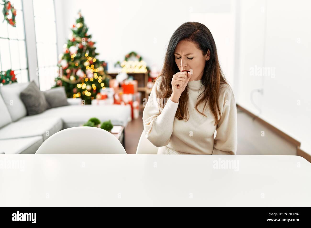 Beautiful caucasian woman sitting on the table by christmas tree feeling unwell and coughing as