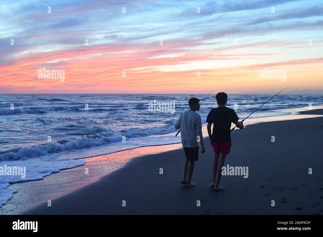 Rear view of two teenage boys walking on the water's edge of the sandy beach with fishing rods at sunset, Marina di Castagneto Carducci, Tuscany Stock Photo