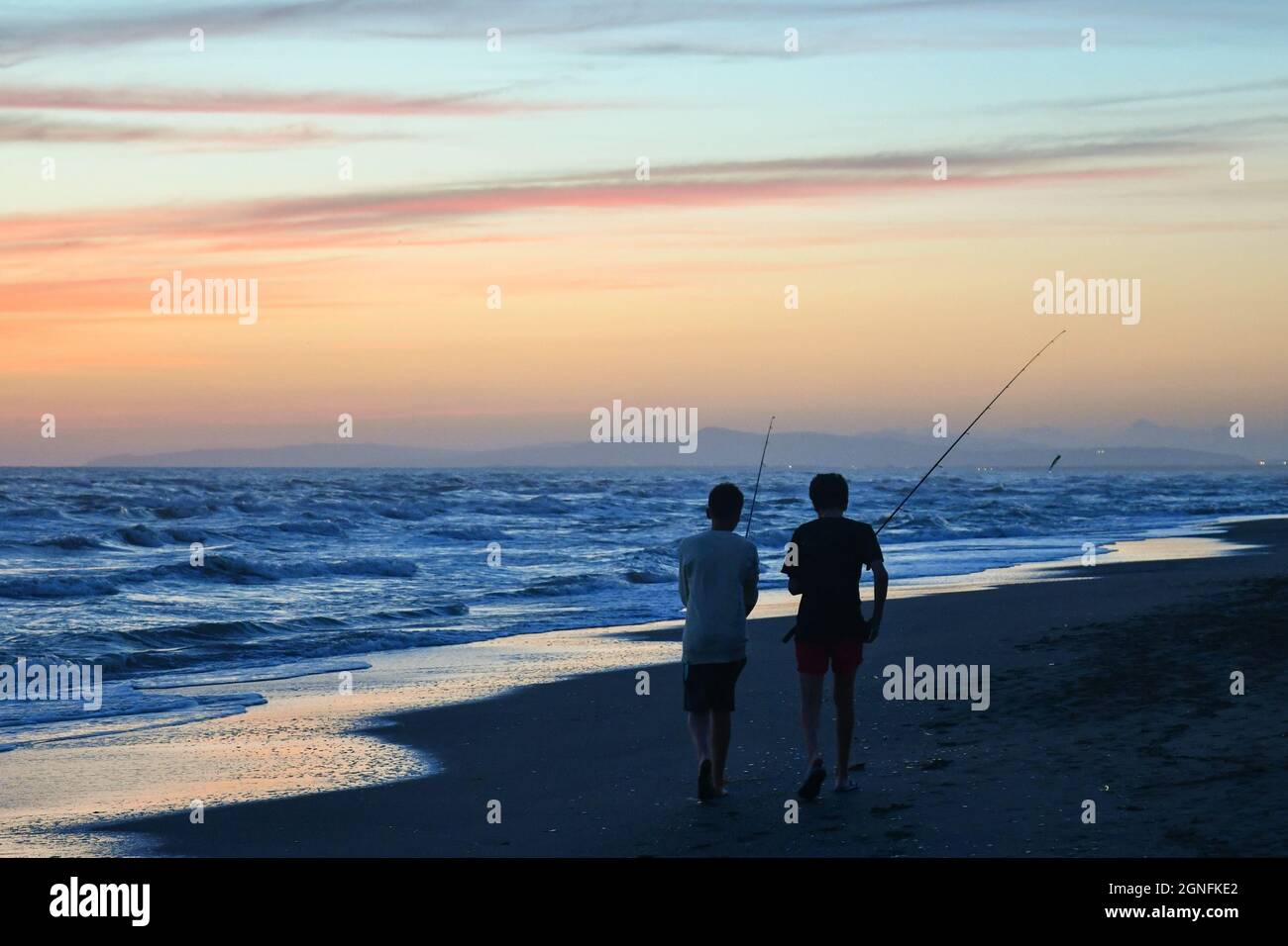 Rear view of two teenage boys walking on the water's edge of the sandy beach with fishing rods at sunset, Marina di Castagneto Carducci, Tuscany Stock Photo