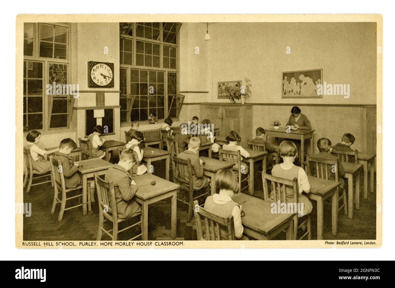 1930's era by Bedford Lemere & Co.  postcard of primary school children, sitting at desks with knitting wool, with teacher, Hope Morley House Classroom, large clock on the wall, Russell Hill School - a boarding school,  Purley, London, England, U.K. Stock Photo