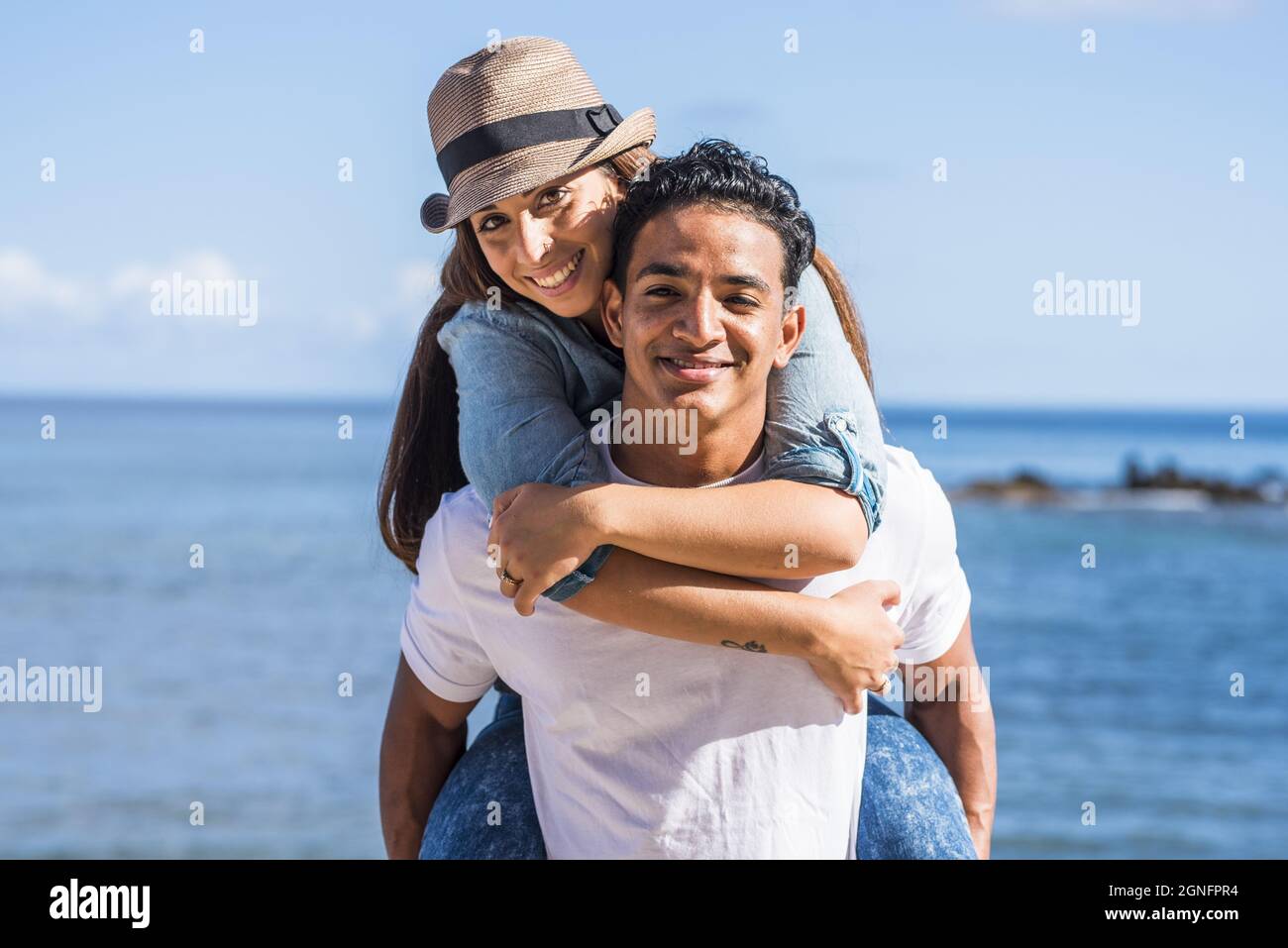 Carefree couple spending leisure time together outdoors, man giving piggyback ride to beautiful girlfriend in front of seascape during summer holiday. Stock Photo